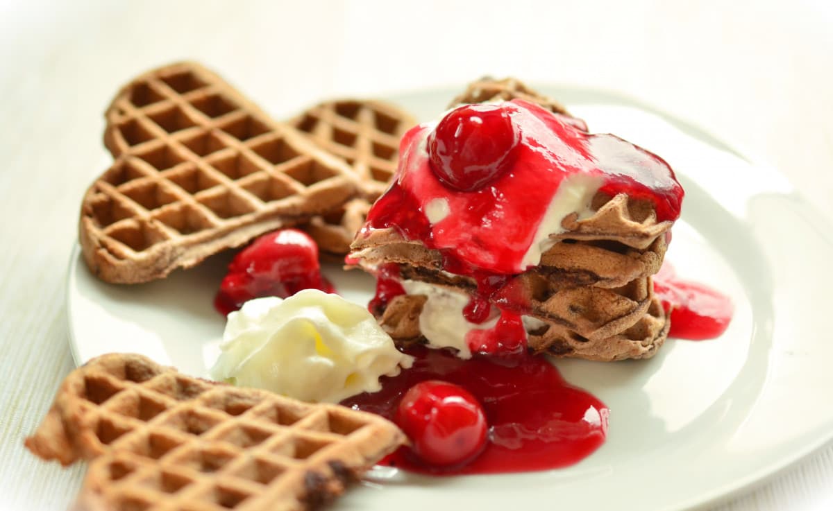 a plate of heart shaped waffles in a stack with cream, sauce, and cherries